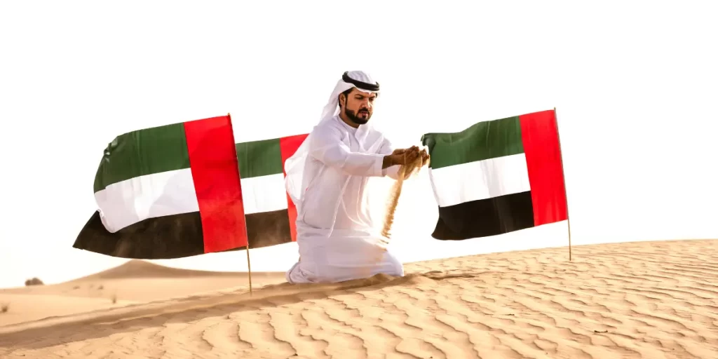 Emirati man in traditional attire celebrating UAE National Day in the desert, surrounded by UAE flags